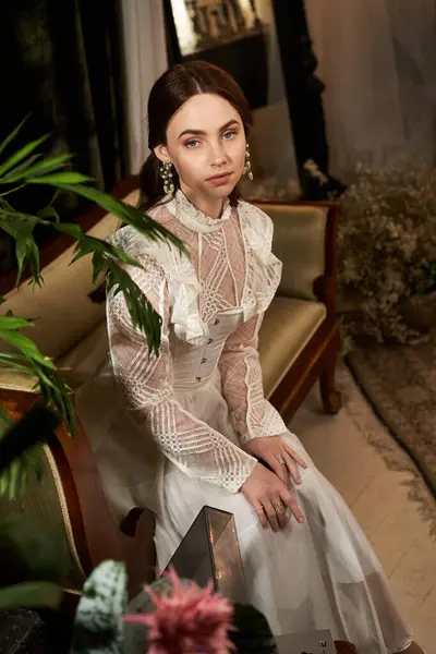 A beautiful young woman in an exquisite vintage dress sits elegantly in a cozy, stylish room. — Stock Photo