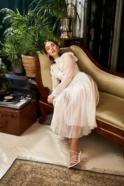 A graceful young woman in an elegant dress sits on a vintage couch amid lush greenery. — Stock Photo