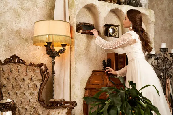 Uma jovem mulher em um vestido vintage decora elegantemente uma prateleira em uma sala quente e convidativa. — Fotografia de Stock