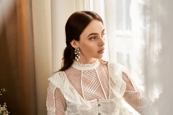 A beautiful young woman dressed in vintage attire gazes thoughtfully through a light-filled window. — Stock Photo