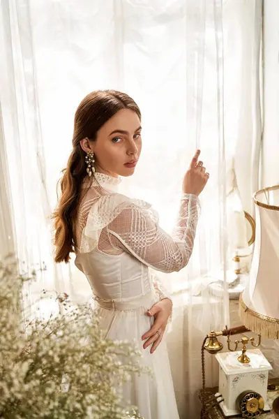 A beautiful young woman in a vintage dress stands by the window, gently touching the sheer curtain. — Stock Photo