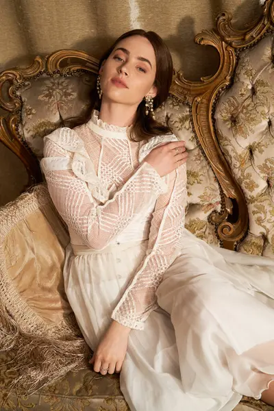 A beautiful young woman in a vintage dress poses gracefully on an ornate sofa, exuding elegance. — Stock Photo