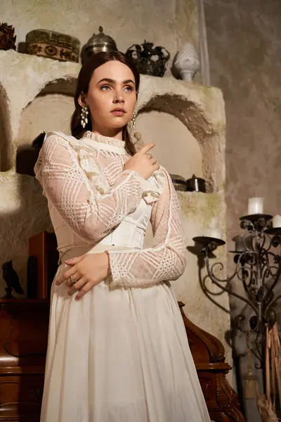 A lovely young woman in a vintage dress stands gracefully among antique decor and soft lighting. — Stock Photo