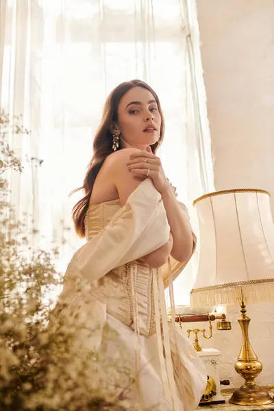 A poised young woman in an elegant vintage gown stands softly lit. — Stock Photo