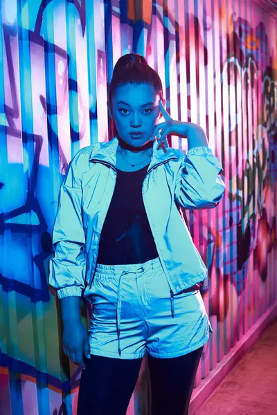 Beautiful woman in shiny jacket and shorts poses artistically in a graffiti filled corridor. — Stock Photo