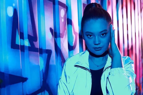 A beautiful woman in a stylish outfit strikes a confident pose near colorful graffiti at night. — Stock Photo