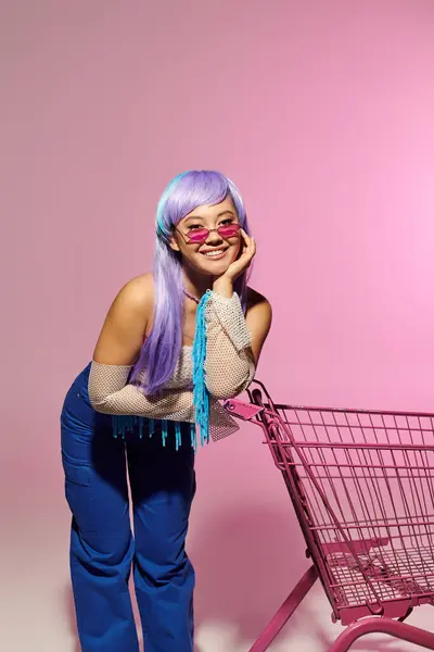 Fashionable woman with purple hair playfully leans on a bright pink shopping cart while smiling. — Stock Photo