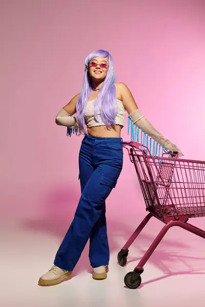 A fashionable woman showcases her unique style beside a bright pink backdrop and shopping cart. — Stock Photo