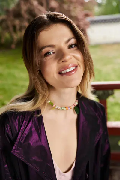 Smiling woman in purple jacket and beads by garden fence. — стоковое фото
