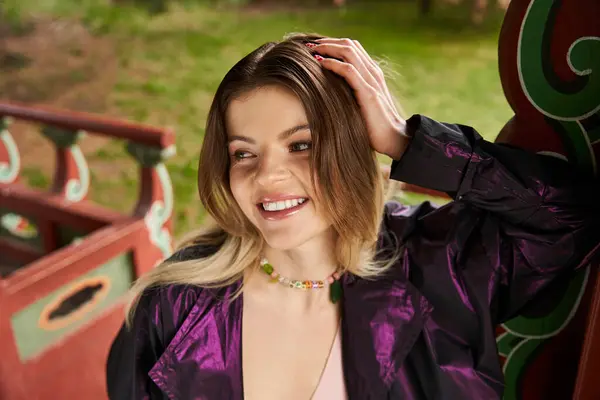 Smiling girl in vivid Park, necklace bright, touches hair, gazing afar. — Stock Photo