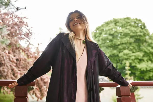 Woman in fancy coat smiles, leaning on veranda railing. Green trees visible. — Stock Photo