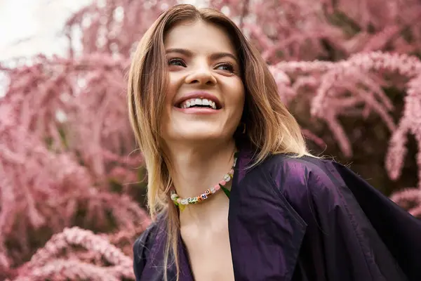 Eine junge Frau mit einem strahlenden Lächeln trägt eine bunte Halskette, während sie von rosa Blüten umgeben ist. — Stockfoto