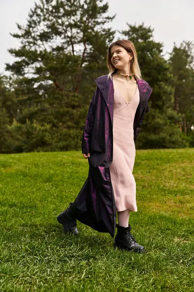 Woman in iridescent coat and pink dress walks green field. — Stock Photo