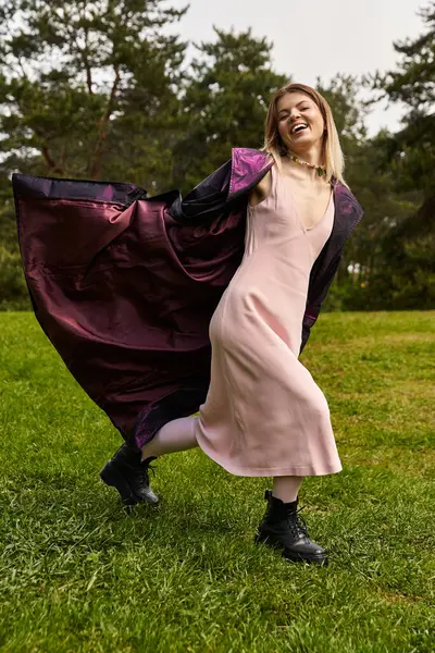 A pretty young woman in a light pink dress and cloak runs cheerfully across a grassy meadow. — Stock Photo