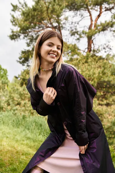 A pretty young woman in a dark coat smiles serenely as she stands outside in front of green trees. — Stock Photo