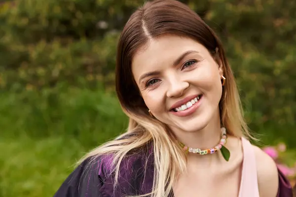 Mujer joven con una sonrisa brillante, se encuentra al aire libre con follaje verde detrás de ella. - foto de stock