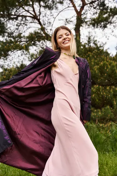 Una joven con un vestido rosa sonríe con alegría, su abrigo ondeando en la brisa en una zona boscosa. - foto de stock