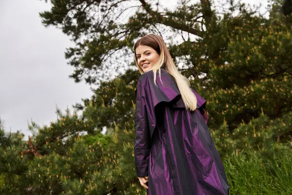 Purple coat, soft smile, young woman glances back in green. — Stock Photo