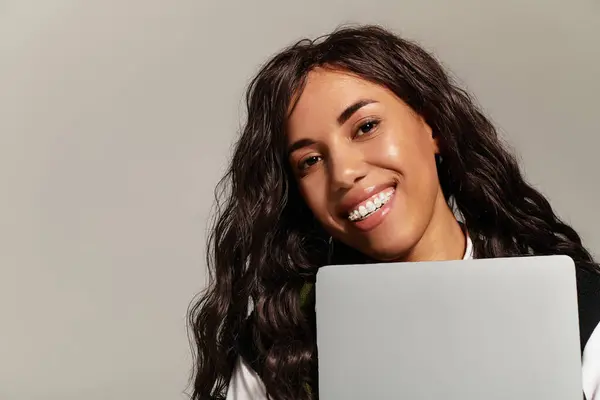 A young woman smiles brightly while holding a laptop against her chest in a studio setting. — стоковое фото