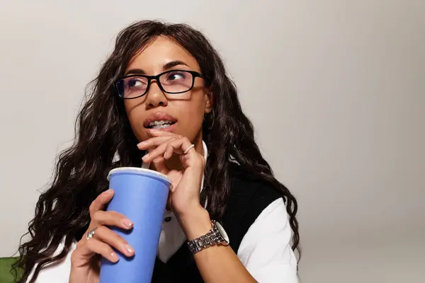 Una bella giovane donna con gli occhiali tiene un drink, persa nel pensiero in una posizione elegante. — Foto stock