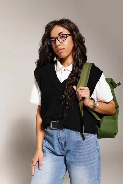 A young student wearing glasses and carrying a backpack poses. She wears a sweater vest and jeans. — Stock Photo