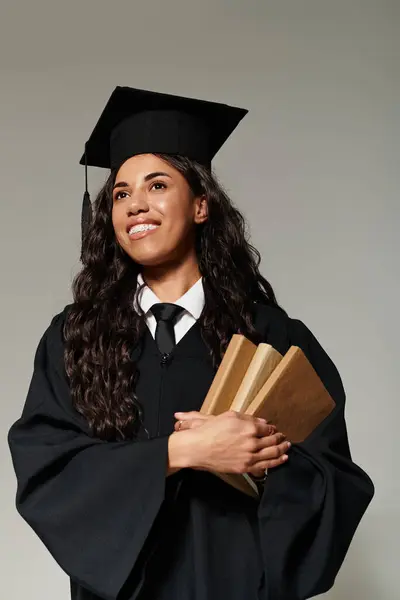 Mujer joven en traje de graduación vigas con orgullo mientras sostiene sus libros, mostrando su éxito. - foto de stock