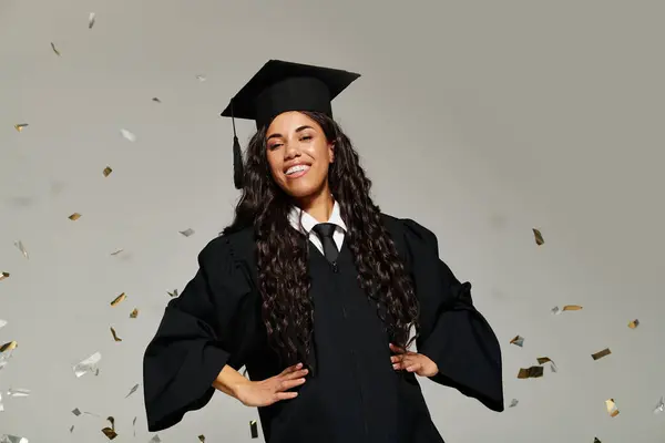 Young woman in graduation cap and gown smiles brightly as confetti falls around her in a studio. — Stock Photo