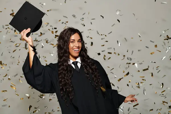 A young woman in graduation attire joyfully throws her cap while surrounded by shimmering confetti. — Stock Photo