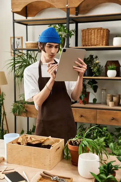Joven guapo que atiende a las plantas en un estudio bien iluminado, centrado en su tarea y usando una tableta. - foto de stock