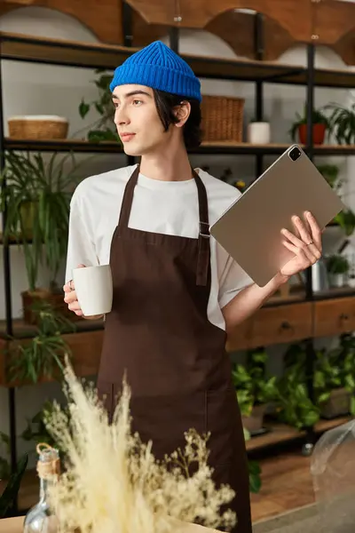 Young man in a brown apron and blue beanie is carefully transplanting plants while holding a tablet. — Stock Photo