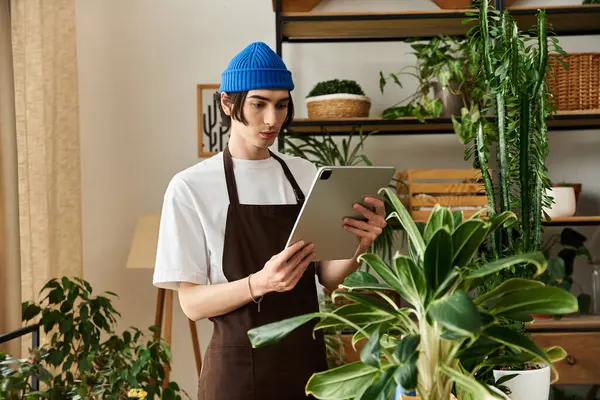 A young man in a blue beanie monitors plant growth with a tablet in a bright studio. — Stock Photo