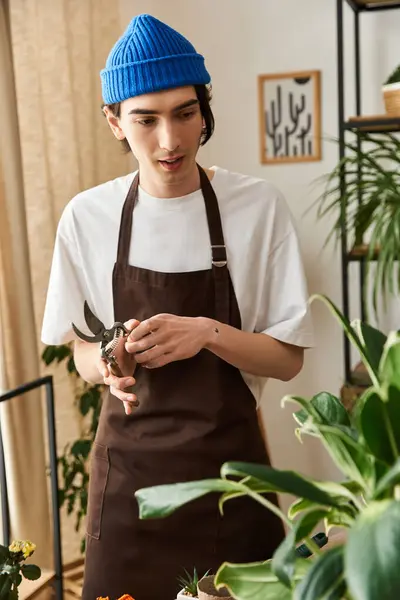 A skilled young man dons a beanie and apron while transplanting vibrant plants in a cozy studio. — Stock Photo