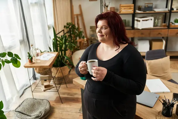 Elegant woman holds a cup, surrounded by natural light and artistic tools in her studio space. — Stock Photo