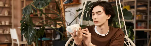 Hombre guapo en el estudio de cerámica examina cuidadosamente su creación de cerámica única entre las plantas. - foto de stock