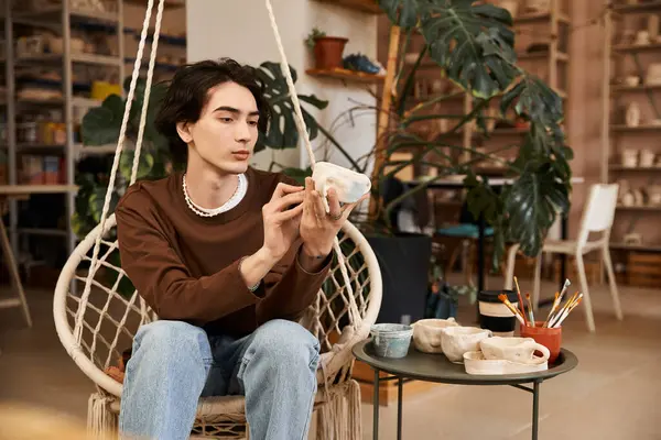 A stylish young man carefully examines his pottery creation in a vibrant studio brimming with art. — Stock Photo