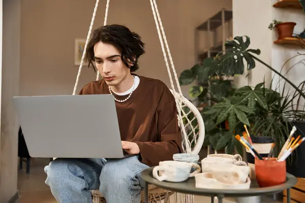El hombre con estilo se centra en su portátil mientras está rodeado de cerámica y plantas en el estudio. - foto de stock