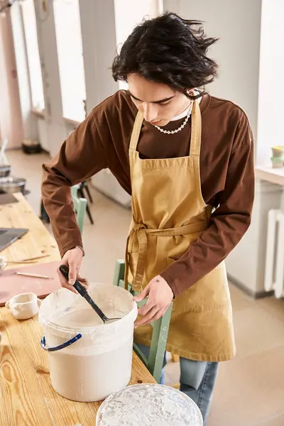 Handsome man in an apron mixes materials in a bright pottery studio dedicated to art and creativity. — Stock Photo