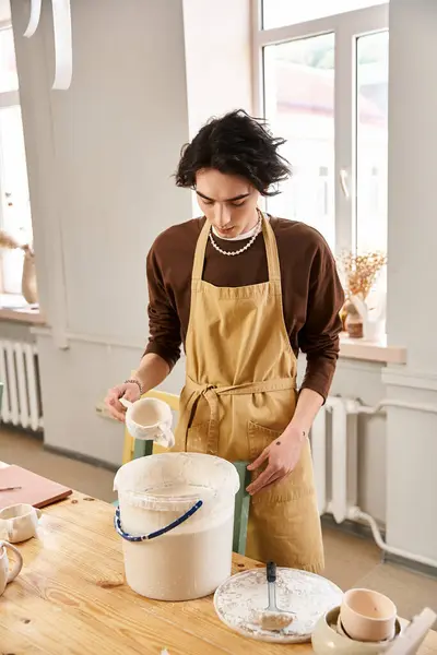 A focused and stylish man pours clay from a container while surrounded by various pottery tools. — Stock Photo