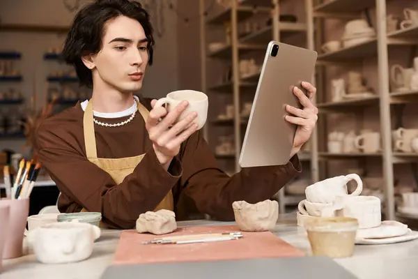 Un hombre en un estudio de cerámica examina una taza de arcilla mientras usa una tableta para la inspiración creativa. - foto de stock