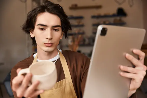 Man in pottery studio holds a cup and focuses on a tablet, merging tradition with tech. — Stock Photo