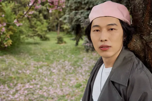 A young asian man stands in a park, wearing a trendy grey outfit, surrounded by beautiful blossoms. — Stock Photo