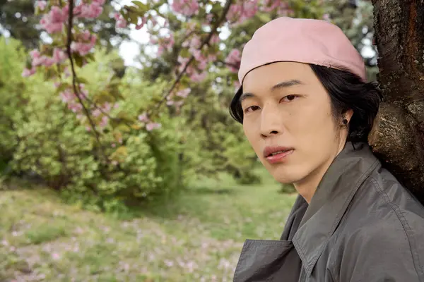 Young man in a trendy grey trench coat relaxes under a blooming tree in the park. — Stock Photo