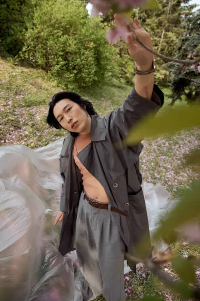 A young asian man showcases his trendy grey outfit while exploring a lush park during spring. — Stock Photo