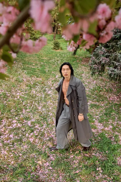 In a vibrant park, a young man flaunts a grey trench coat among spring blossoms. — Stock Photo