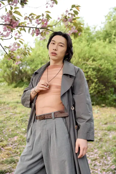 A young Asian man stands confidently in a stylish grey trench coat, embracing nature's beauty. — Stock Photo