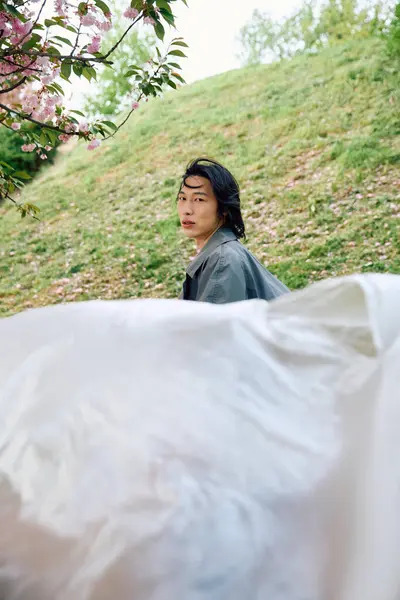 Un jeune asiatique dans un élégant trench coat gris posant avec du polyéthylène dans un parc verdoyant. — Photo de stock