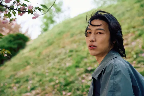In a peaceful park, a young asian man in a grey outfit expresses stylish confidence while relaxing. — Stock Photo