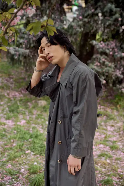 A young asian man in a grey trench coat interacts with nature in a floral park setting. — Stock Photo