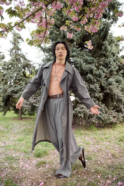 A young Asian man showcases a trendy grey outfit while strolling through a vibrant park in spring. — Stock Photo