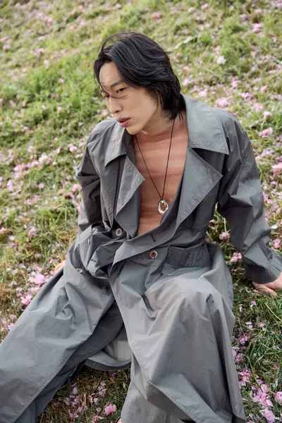 Un joven asiático se pone una elegante gabardina gris, sentada entre delicadas flores en el parque. — Stock Photo